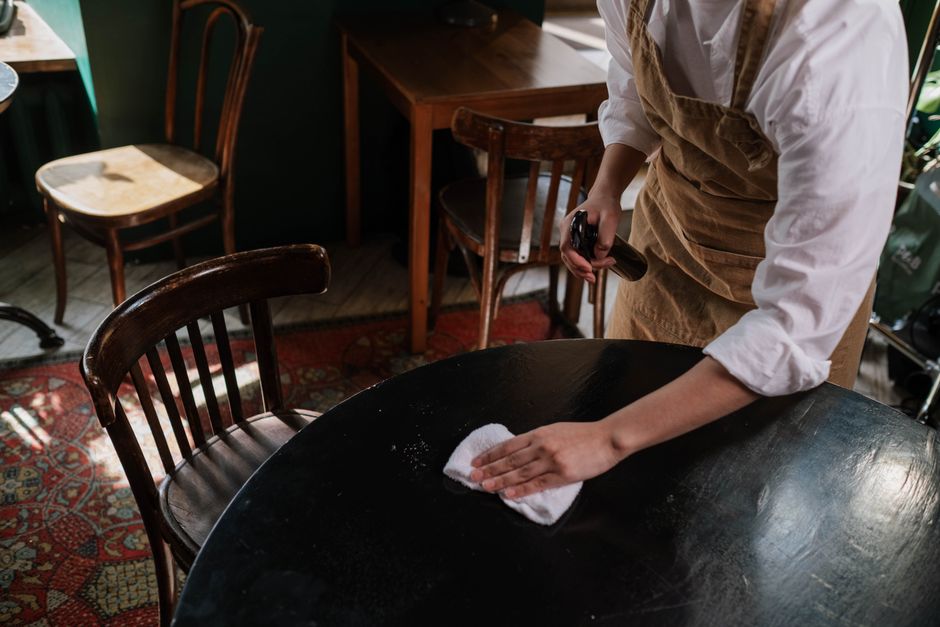 A person wearing a white shirt and brown apron is performing surface cleaning on a dark wooden round table, using a white cloth to wipe the surface. The individual is holding a spray bottle in the oth