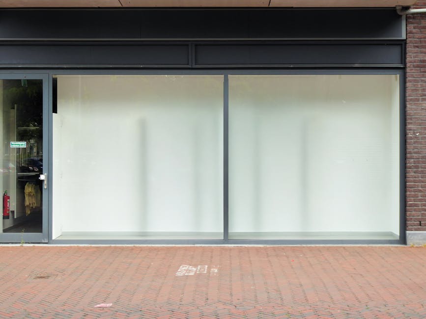 Close-up view of a shopfront on Colliers Wood High Street, featuring a glass display window and metal shutter, with minor dust visible on the glass surface. The window is framed by a blue metal struct